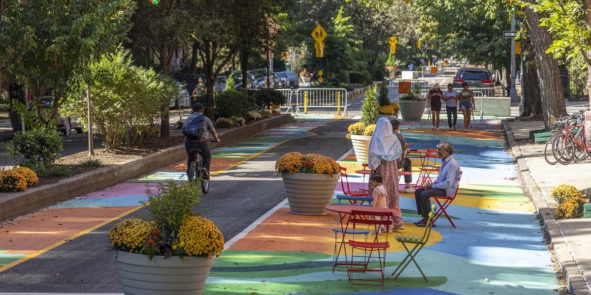 Paseo Park Queens NYC with brightly colored pavement, trees and tables and people bicycling and sitting at tables in chairs.