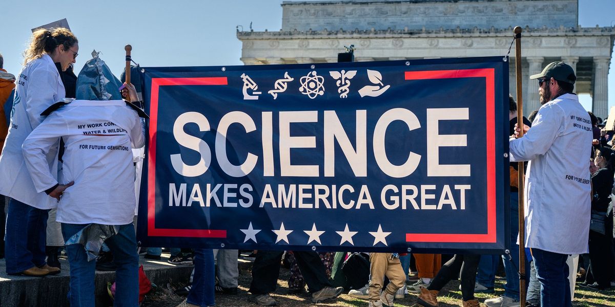 People in white lab coats stand outside with a large blue banner reading "science makes america great."