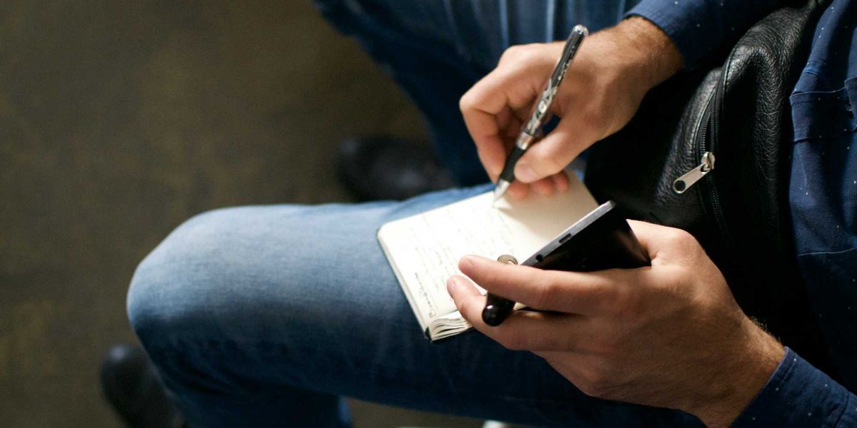 Person holding a cell phone and notepad on their lap while writing.