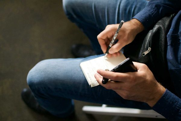Person holding a cell phone and notepad on their lap while writing.