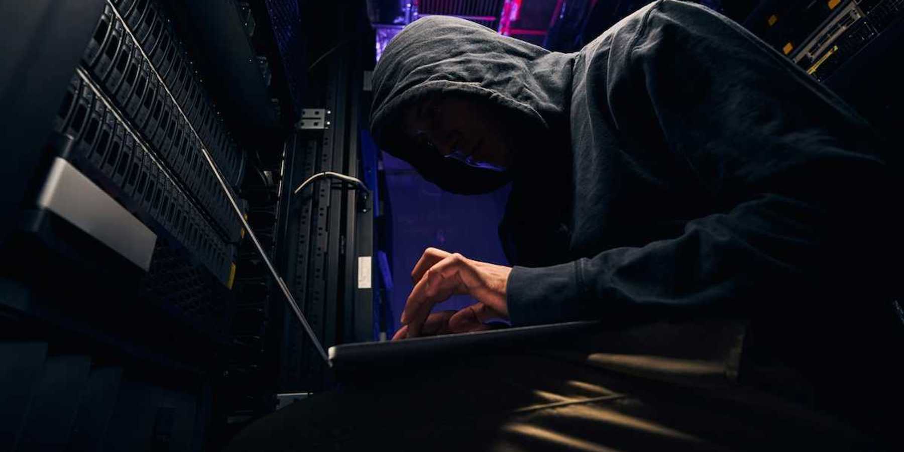 Person in black hoodie, hunched over a keyboard, personification of a shadowy computer hacker.