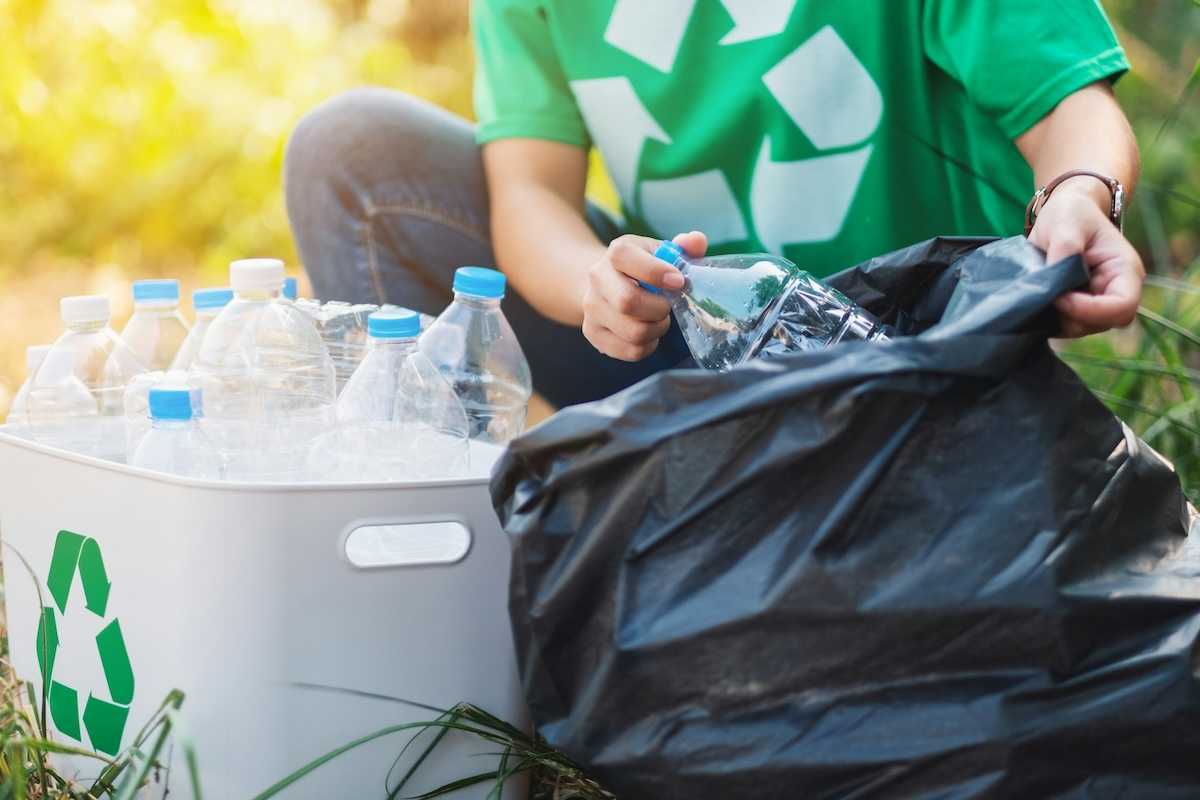 Person in green shirt featuring universal recycling logo sorting plastic boittles from trash bag into recycling container