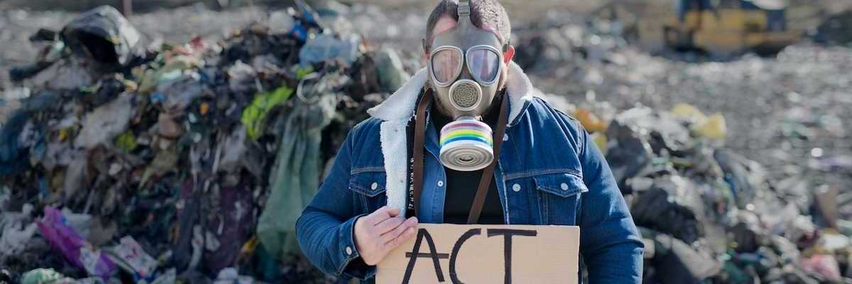 Person standing in an active landfill, wearing respirator and holding a cardboard sign reading "ACT NOW"