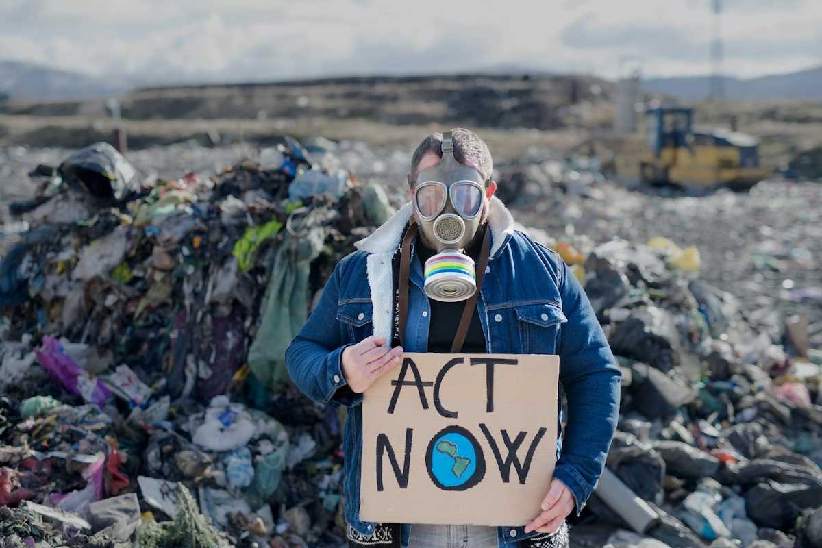 Person standing in an active landfill, wearing respirator and holding a cardboard sign reading "ACT NOW"