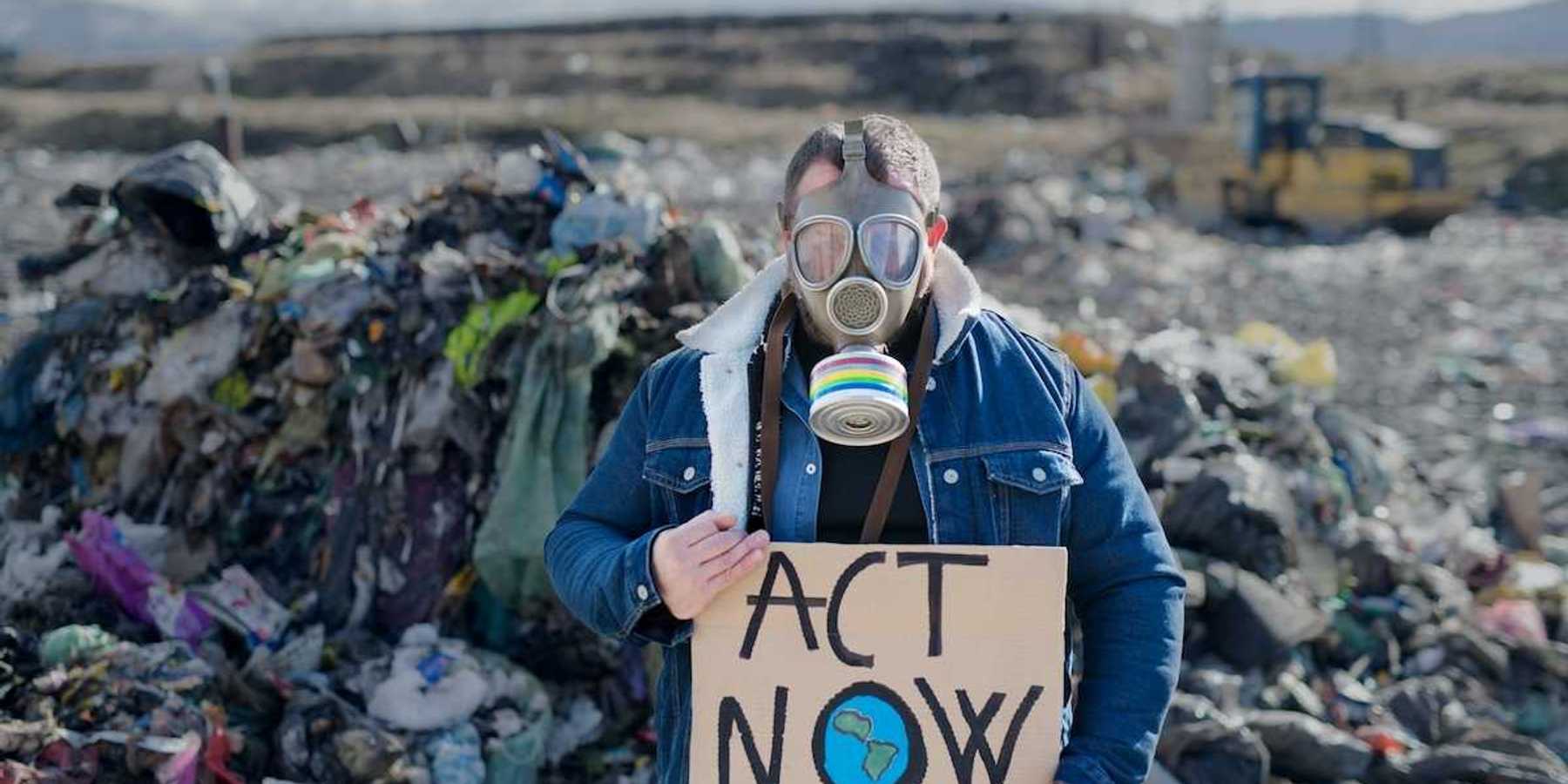 Person standing in an active landfill, wearing respirator and holding a cardboard sign reading "ACT NOW"