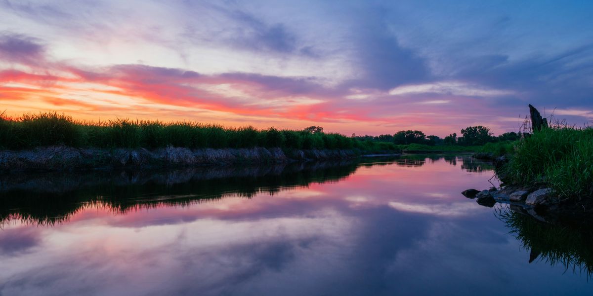photo of body of water surrounded with grass field at sunrise with an orange and pink sky.