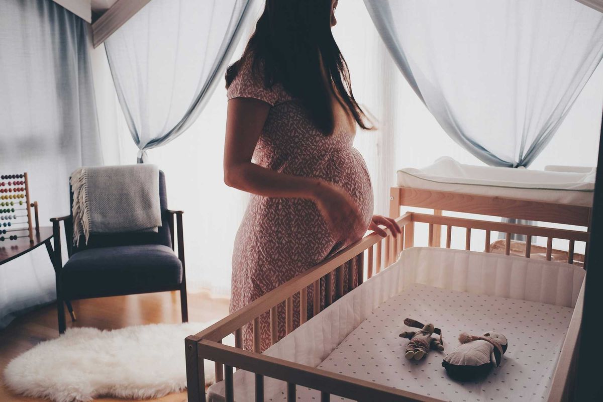 Pregnant woman in white lace sleeveless dress standing beside brown wooden crib.