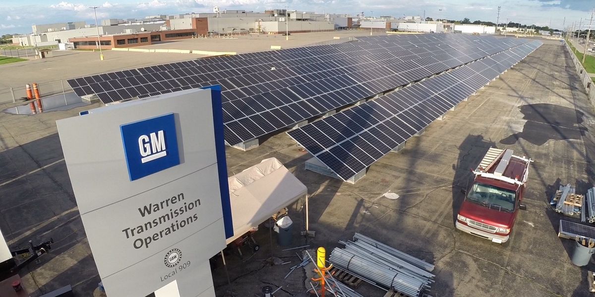 Rows of solar panels in a large parking lot with a sign in the foreground displaying a General Motors logo.