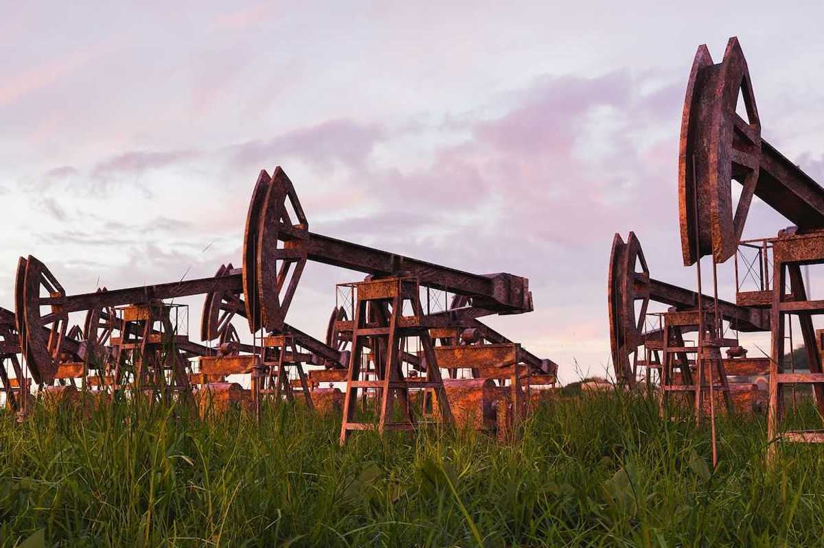 Rusted and abandoned oil pumps with grass growing around it against sunset.