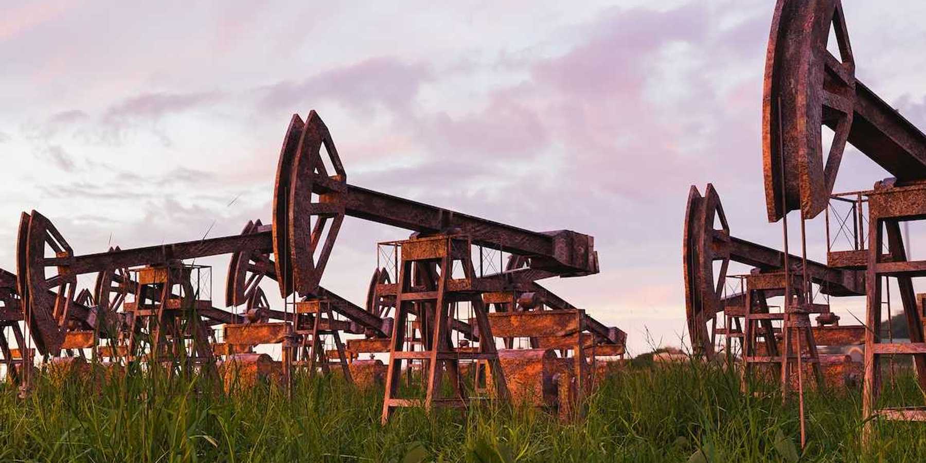 Rusted and abandoned oil pumps with grass growing around it against sunset.