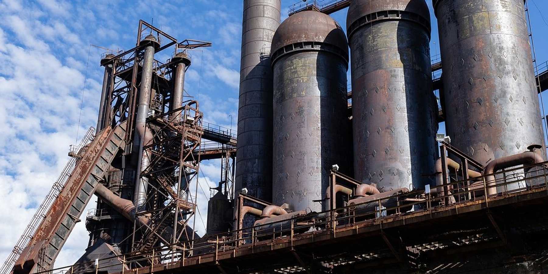 Rusting abandoned steel mill, blast furnaces and smokestacks atop a concrete wall.