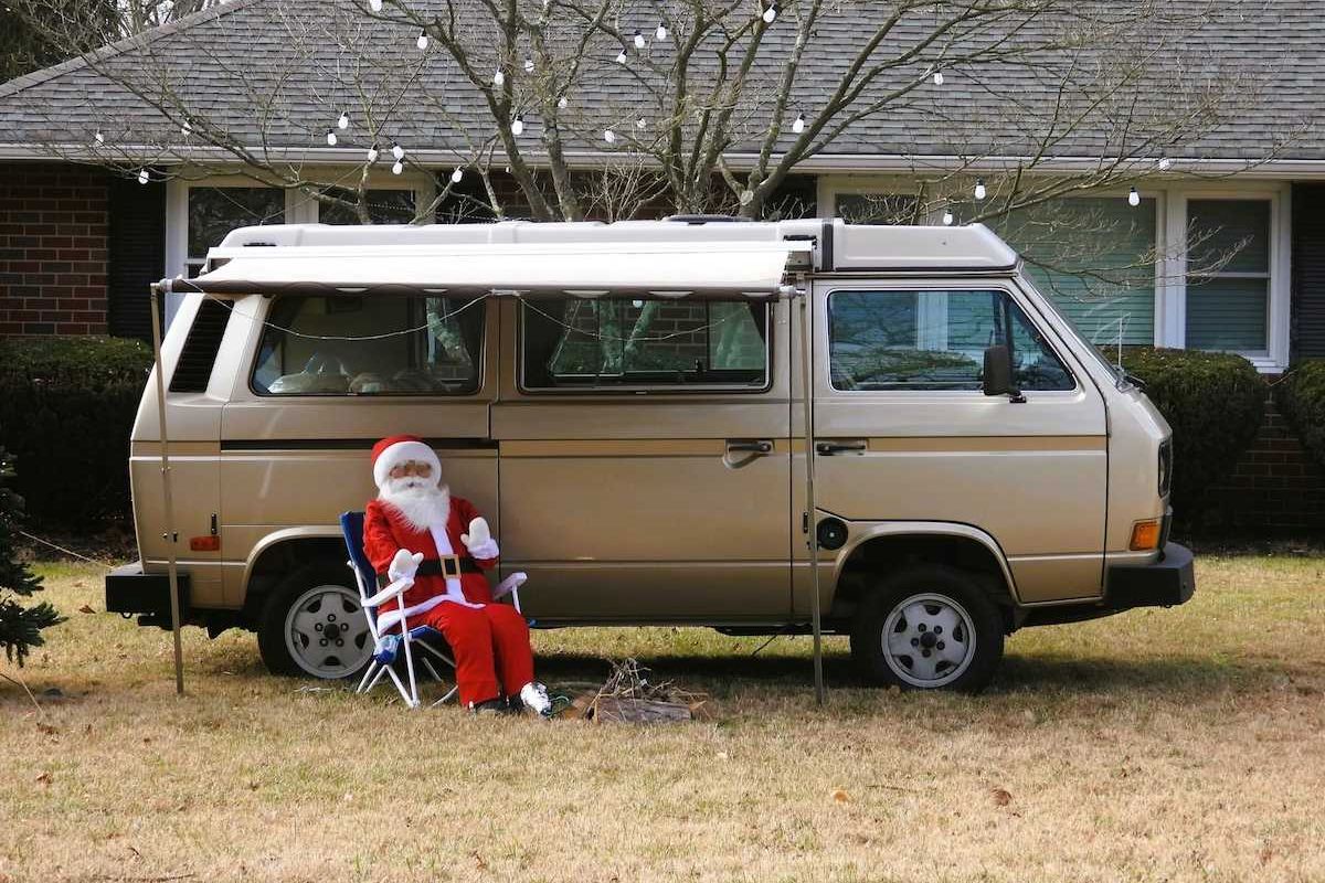 'Santa Claus' sitting in lawn chair adjacent to a camper van on snowless lawn