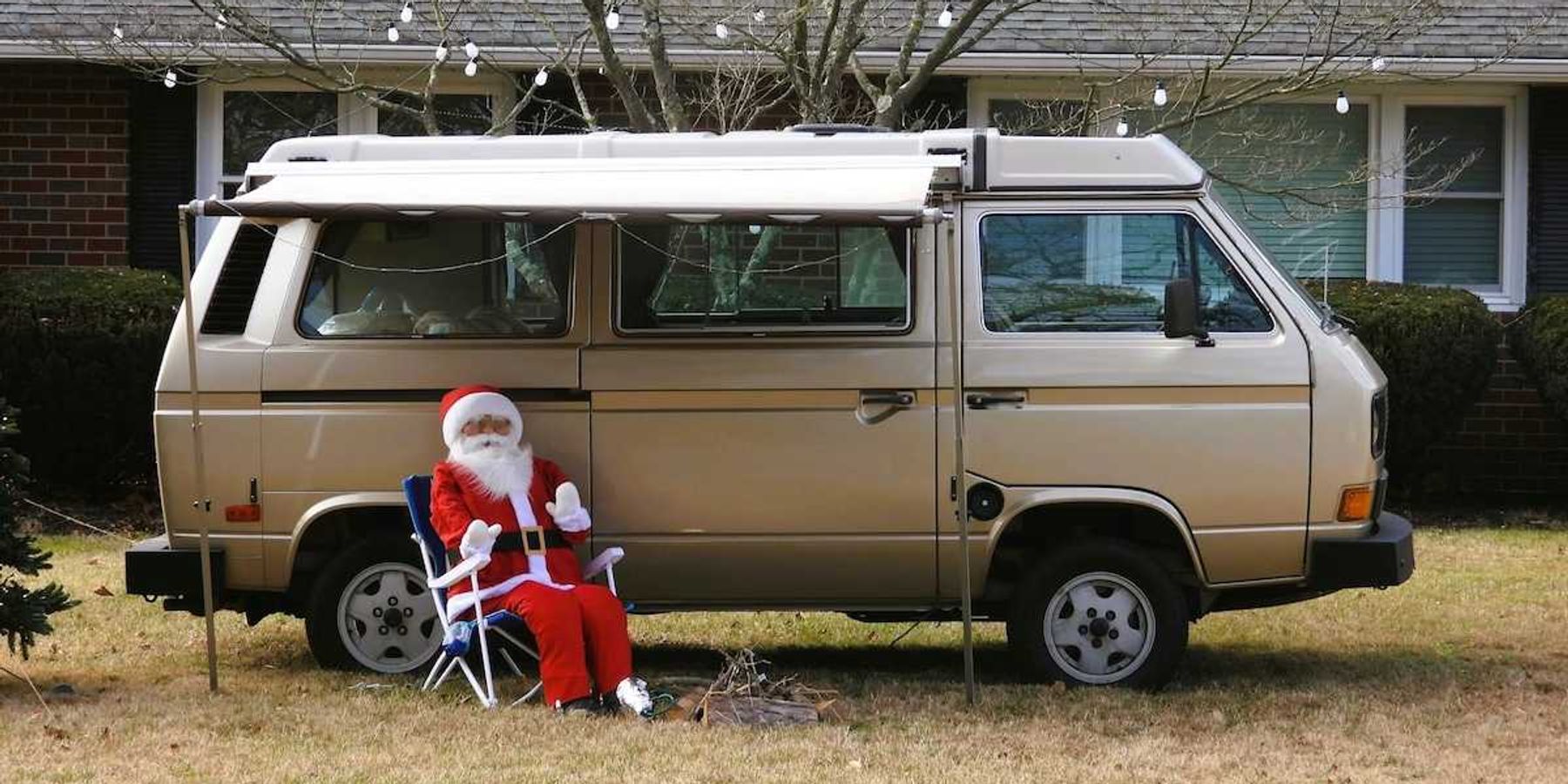 'Santa Claus' sitting in lawn chair adjacent to a camper van on snowless lawn