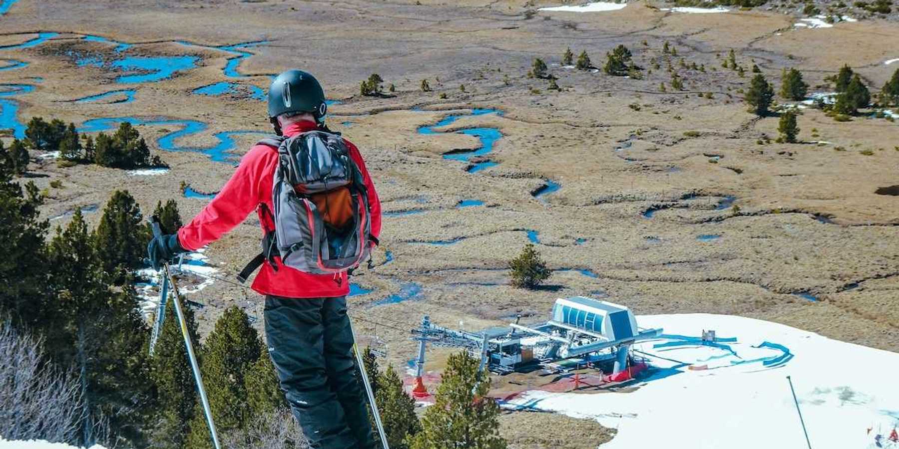 Skier in red jacket poised to descend to lift station. Spotty snow cover.