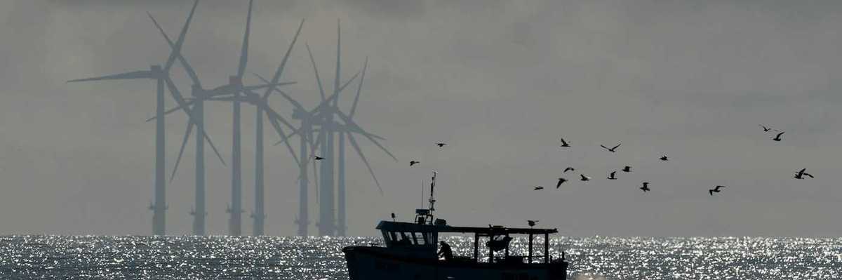 Small fishing boat trailed by sea birds with wind turbines in the background.