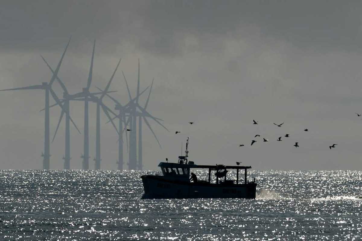 Small fishing boat trailed by sea birds with wind turbines in the background.