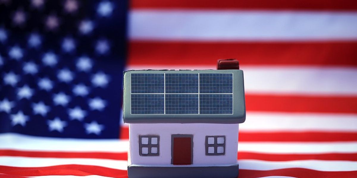 Small white house figurine with solar panels on the roof with an American flag in the background.