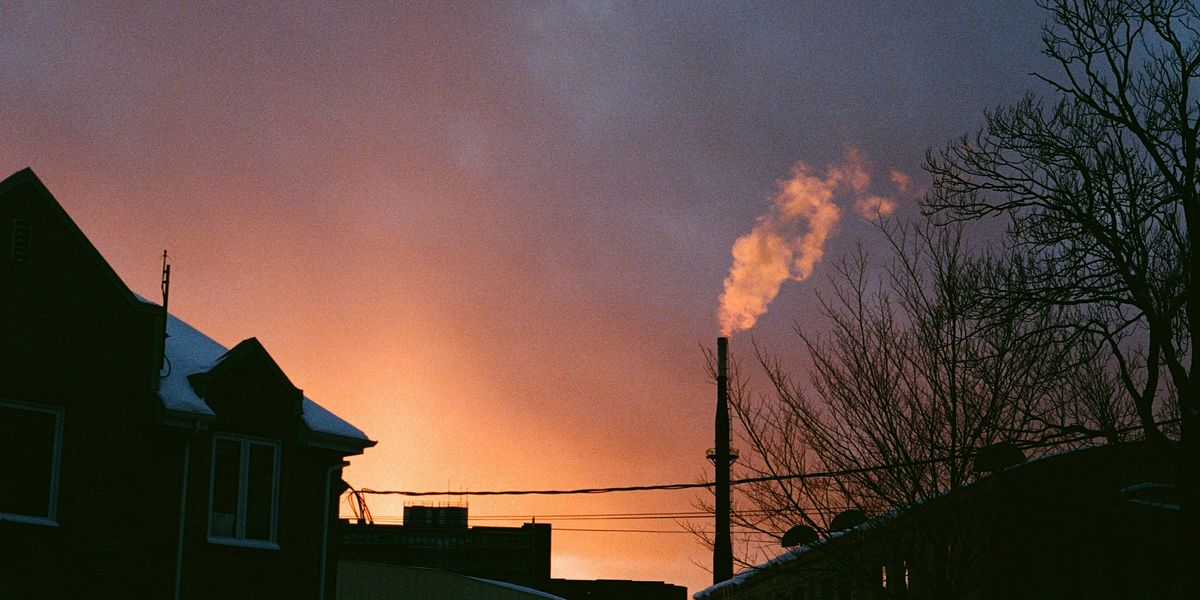 Smoke billows from an industrial chimney at sunset near several homes.