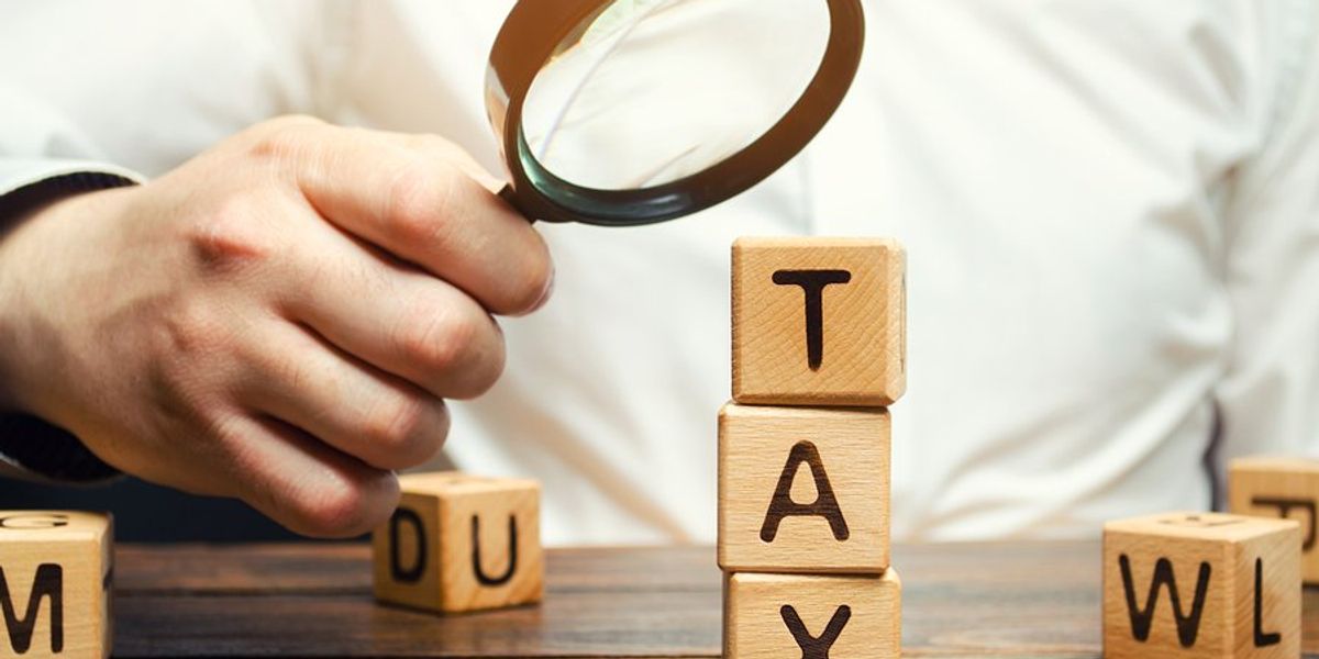 Stack of three wooden blocks with the letters TAX on them with a person holding a magnifying glass on it
