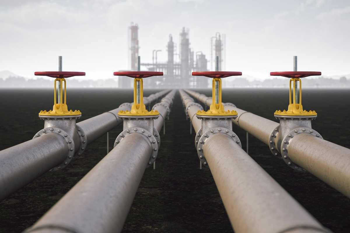 Steel quad-pipeline in the foreground with yellow/red valves, leading to a reinery in the background