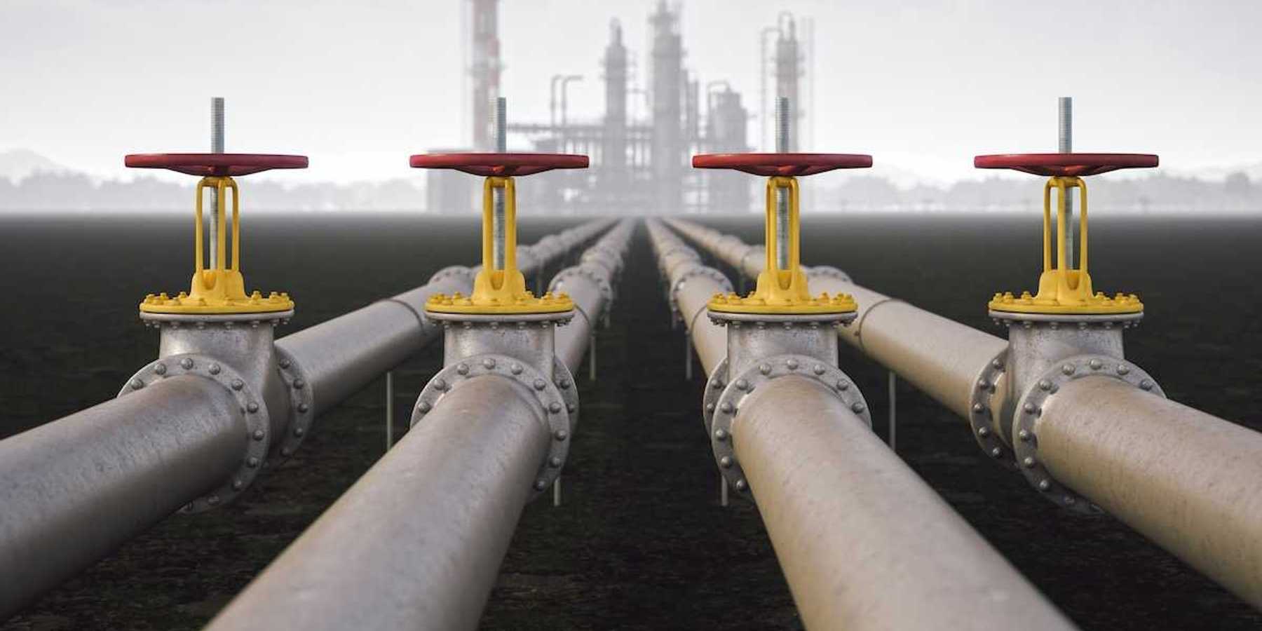 Steel quad-pipeline in the foreground with yellow/red valves, leading to a reinery in the background