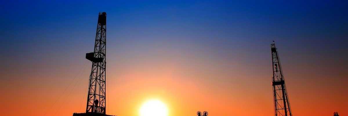 Sun setting over oilfield - drilling rigs and pumpjacks silhouetted against sunset sky.