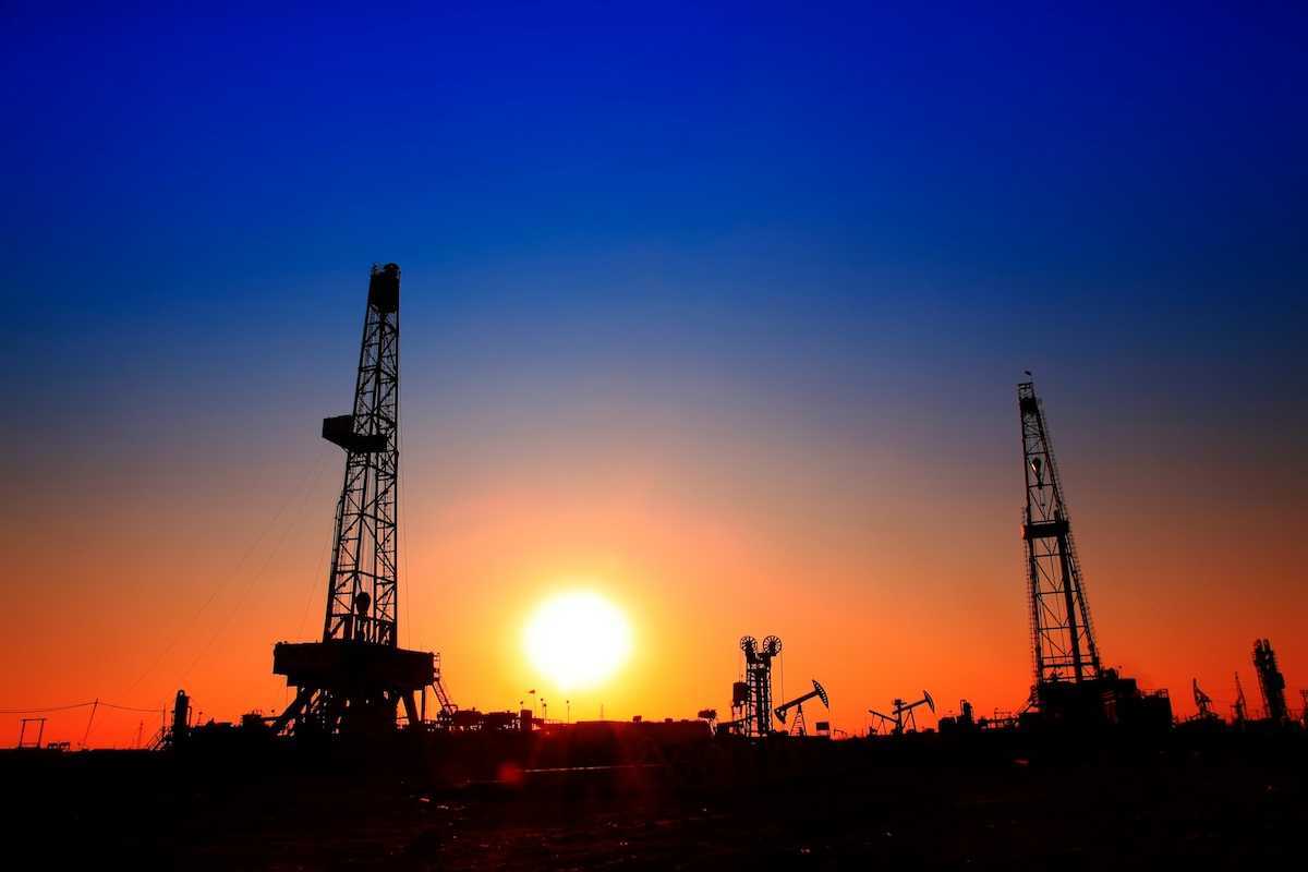 Sun setting over oilfield - drilling rigs and pumpjacks silhouetted against sunset sky.