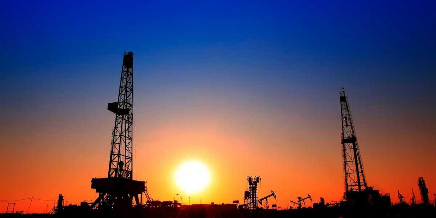 Sun setting over oilfield - drilling rigs and pumpjacks silhouetted against sunset sky.