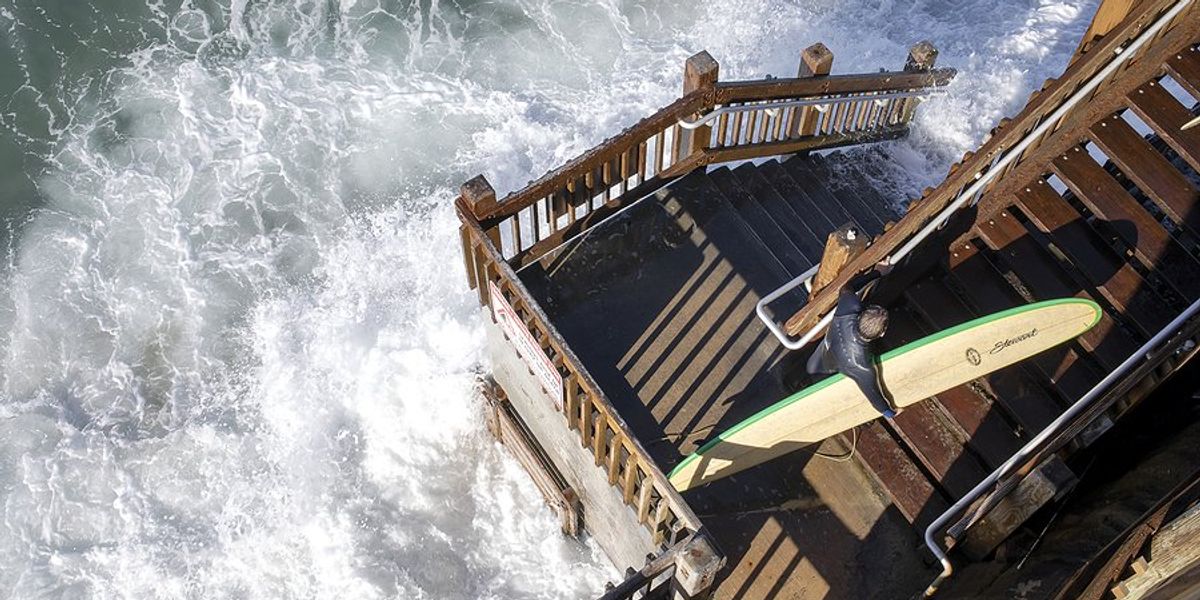 Surfer walking up stairs being inundated by ocean waves.