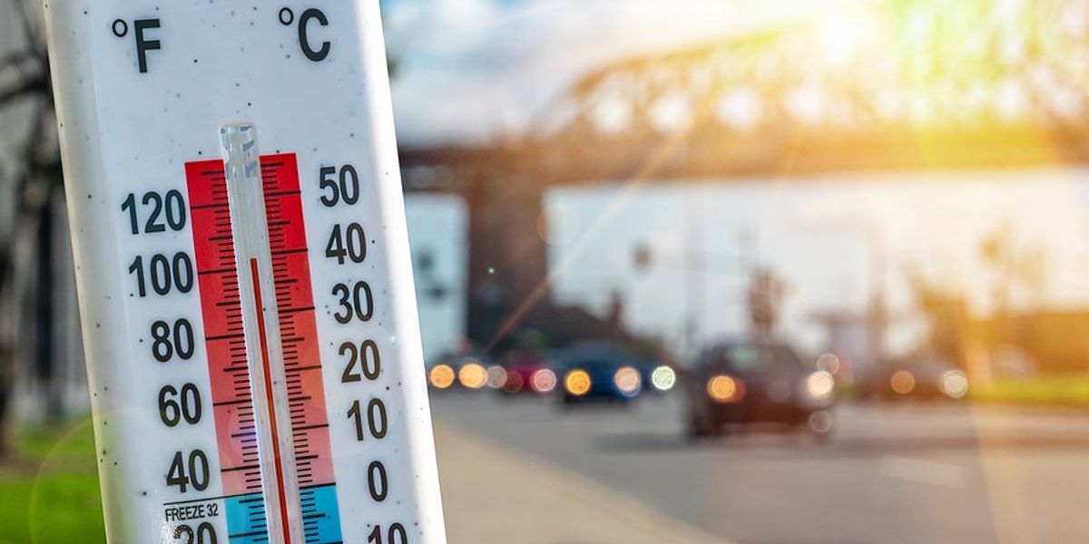 Thermometer in foreground showing hot temperatures in front of an urban landscape of cars and a bridge.