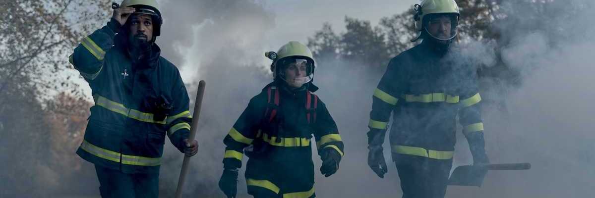 Three firefighters in turnout gear — woman in the center, flanked by two men carrying shovels in smoky air