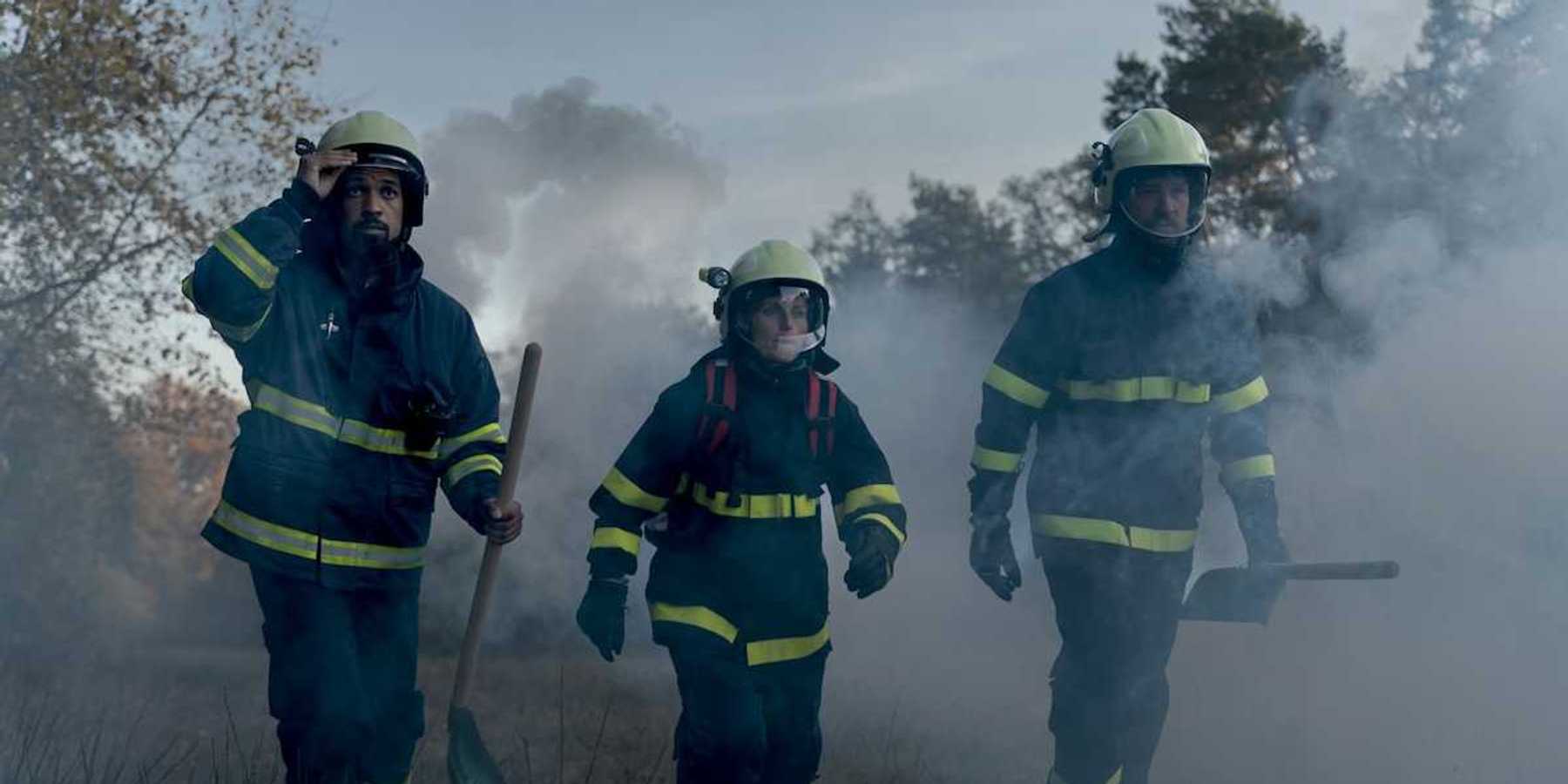 Three firefighters in turnout gear — woman in the center, flanked by two men carrying shovels in smoky air