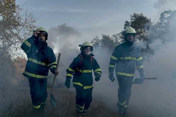Three firefighters in turnout gear — woman in the center, flanked by two men carrying shovels in smoky air