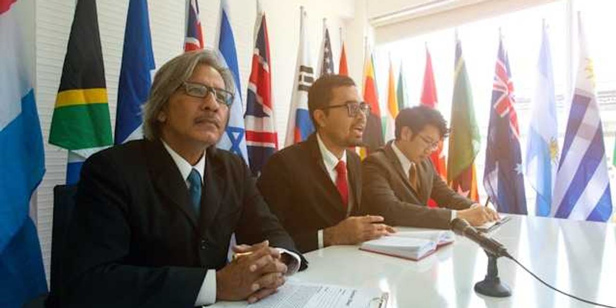 Three men sitting at a conference table with world flags behind them