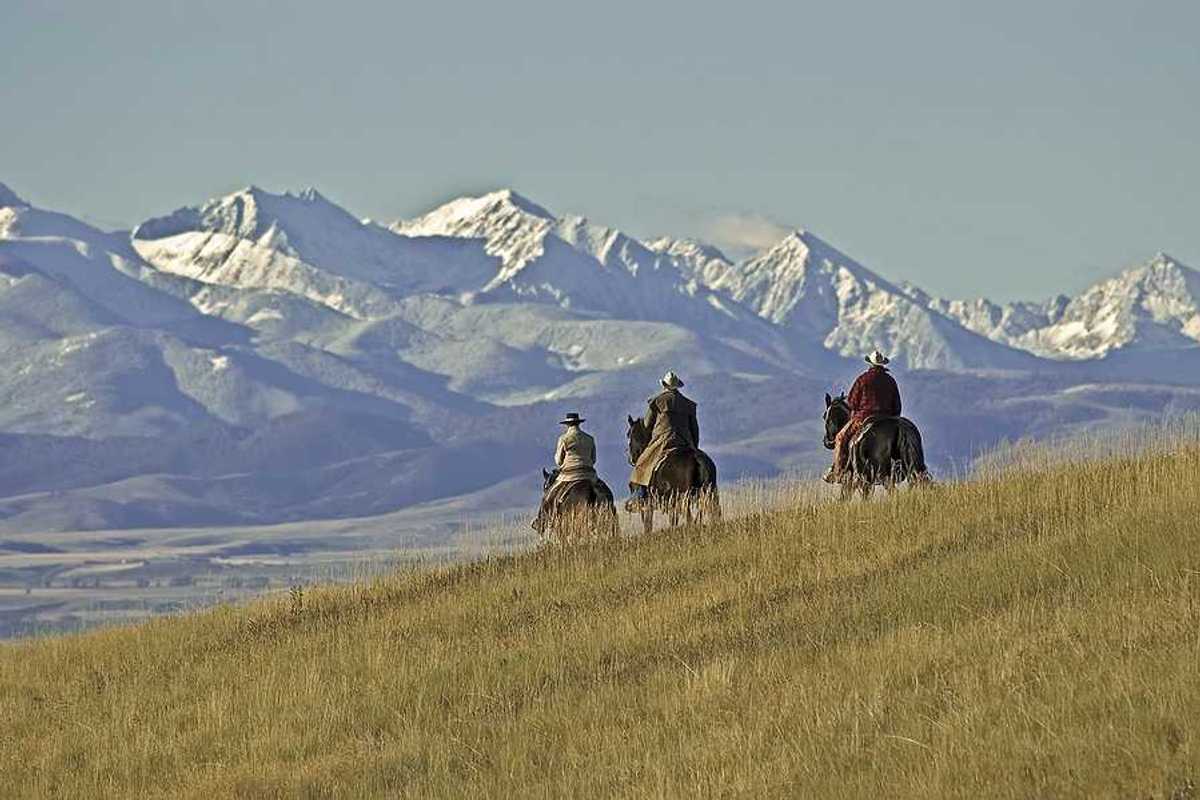 Three people on horses riding over a grassy hill toward mountains