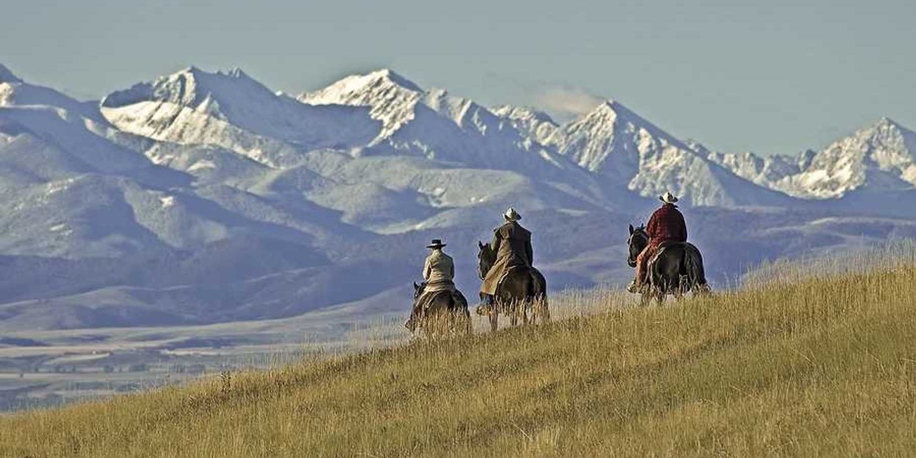 Three people on horses riding over a grassy hill toward mountains