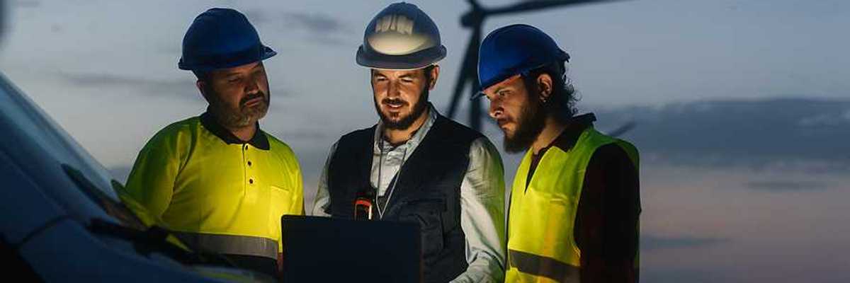 Three renewable energy workers looking at a laptop with a wind turbine in the background