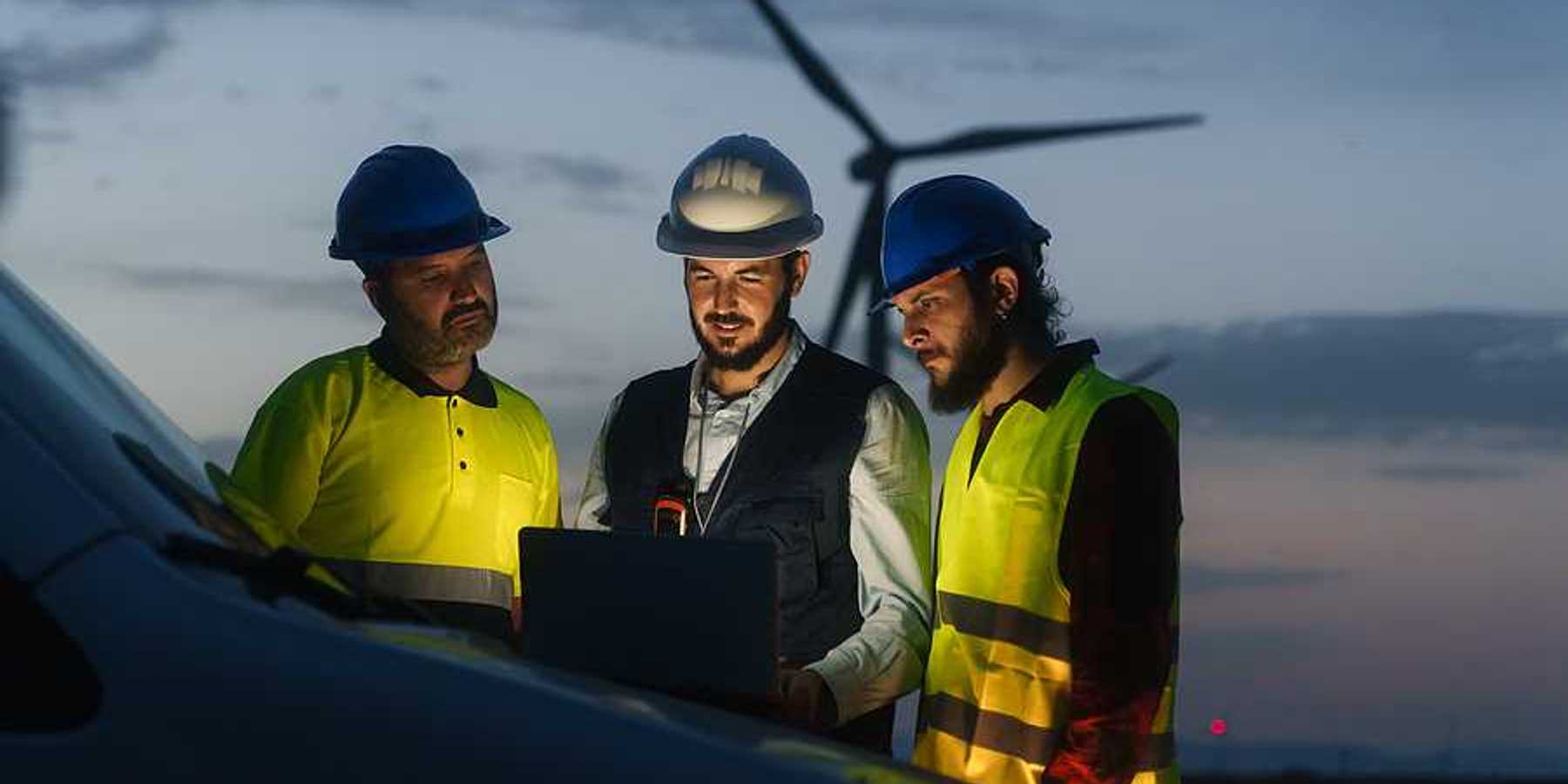 Three renewable energy workers looking at a laptop with a wind turbine in the background