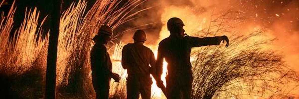 Three silhouetted figures watch a large fire burn in a field