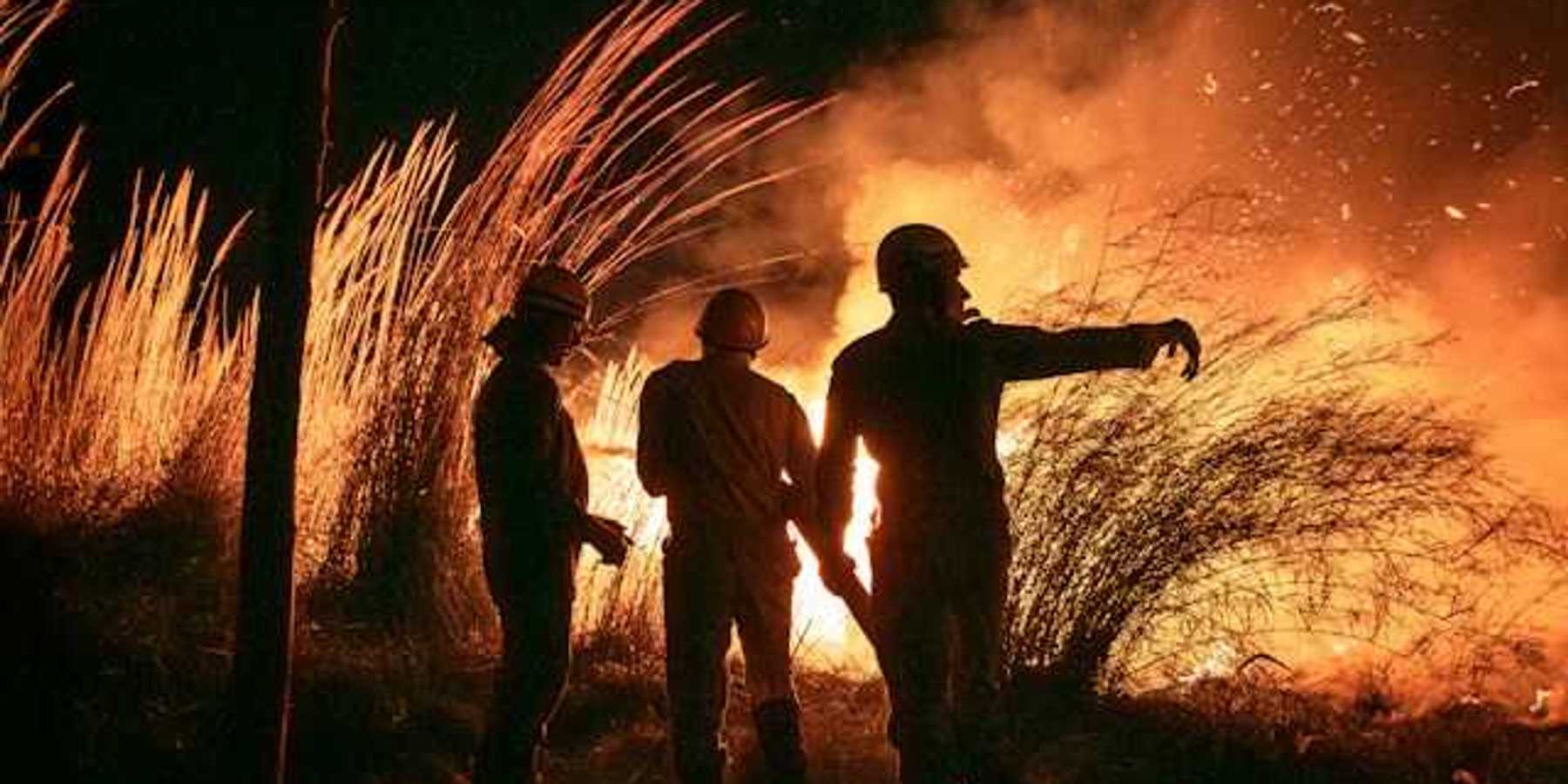 Three silhouetted figures watch a large fire burn in a field
