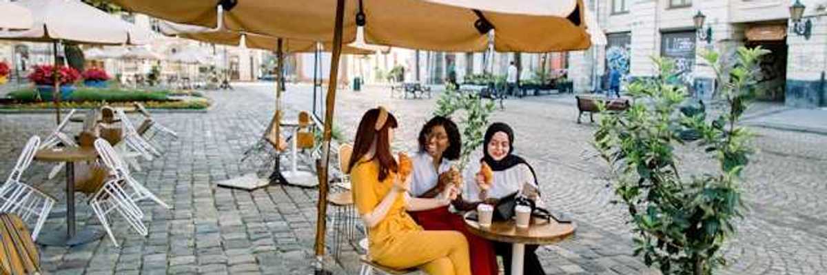Three women sitting under an umbrella in a European city