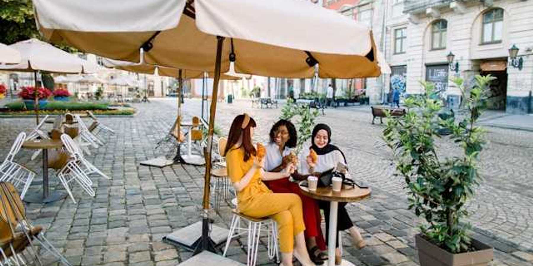 Three women sitting under an umbrella in a European city