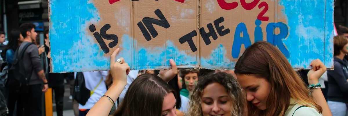 Three young women huddling over a phone or tablet, bearing a handmade sign reading "LOVE CO2 IS IN THE AIR" with the word love crossed out.