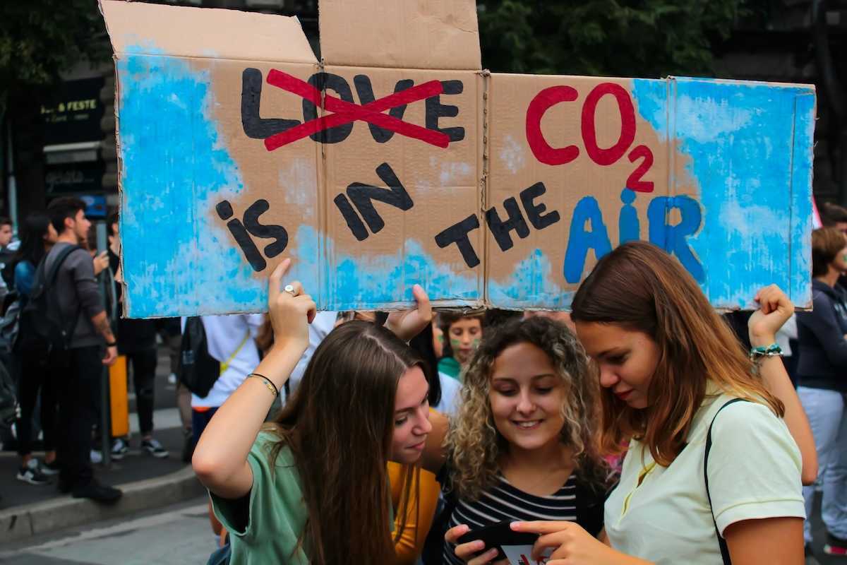 Three young women huddling over a phone or tablet, bearing a handmade sign reading "LOVE CO2 IS IN THE AIR" with the word love crossed out.