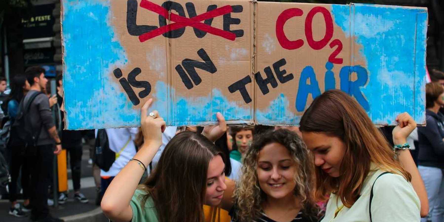 Three young women huddling over a phone or tablet, bearing a handmade sign reading "LOVE CO2 IS IN THE AIR" with the word love crossed out.