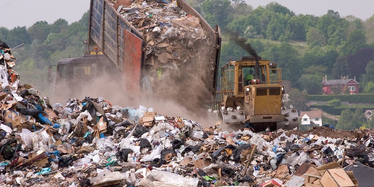 Trash is dumped from a large truck onto an enormous heap of landfill waste, with dust rising and trees in background.