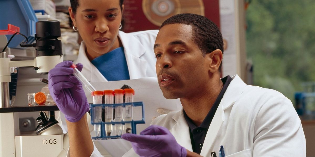 Two Black scientists in lab coats look at a collection of test tubes in a lab.