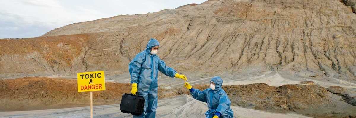 Two field workers in protective garb, sample water near a mining site adjacent to a sign reading Toxic Danger