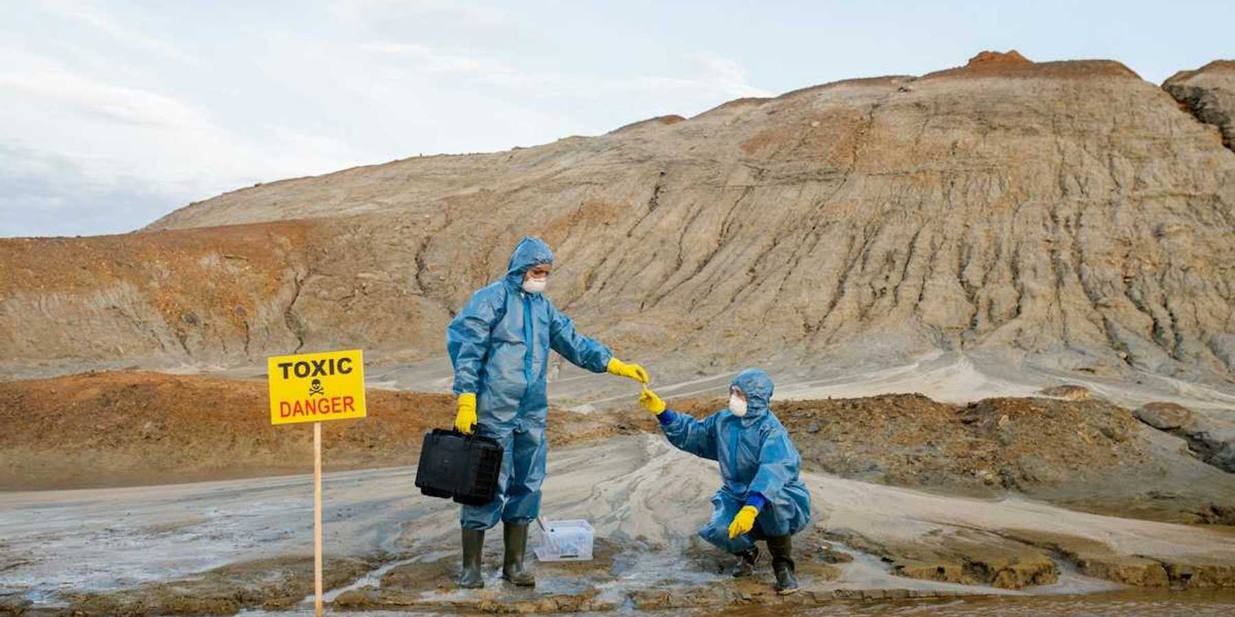 Two field workers in protective garb, sample water near a mining site adjacent to a sign reading Toxic Danger