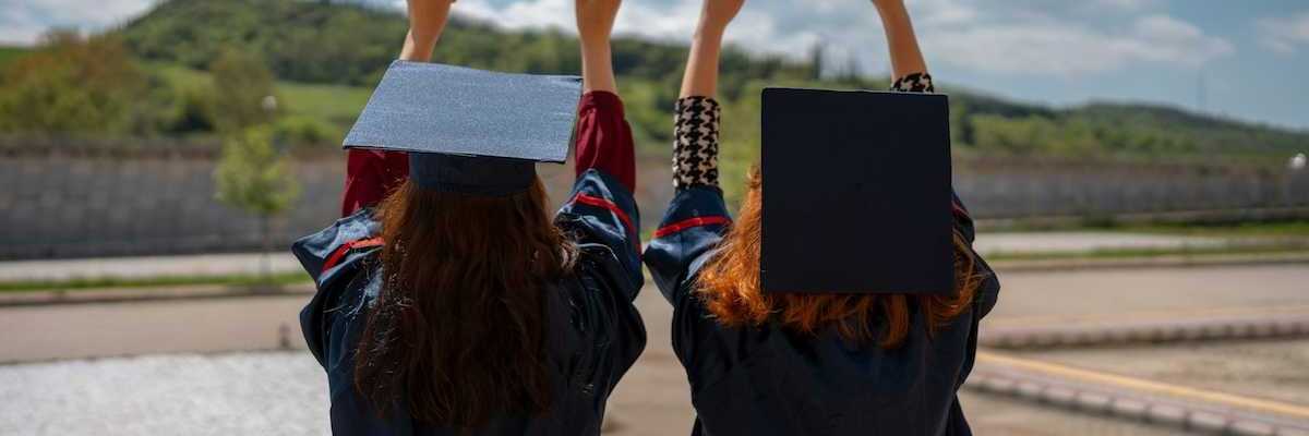 Two graduates with backs to camera holding their rolled-up diplomas above their heads, against the sky
